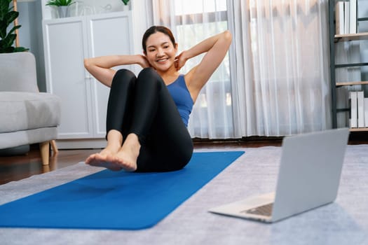 Asian woman in sportswear doing crunch on exercising mat as home workout training routine. Attractive girl engage in her pursuit of healthy lifestyle with online exercise training video. Vigorous