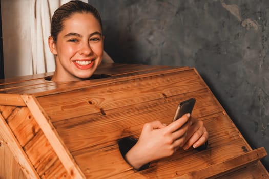 Pretty caucasian girl looking at camera while playing her mobile phone at sauna room. Attractive female with beautiful skin looking at camera during using wooden sauna cabinet. Tranquility.