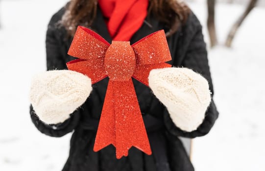 A woman in a black coat and red scarf holds a large red shiny bow in a snowy winter scene. Christmas and New Year concept