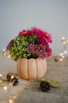 Autumn composition with a bouquet of flowers in a vase in the form of an orange pumpkin. Autumn room decor. Garland with warm light. Bokeh. Thanksgiving Day. Festive composition. Vertical photo.
