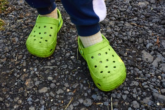 Photo children feet in garden green crocs against the background of a gray road. Life in the village. Comfortable shoes with holes.