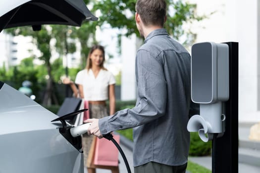 Young couple travel with EV electric car charging in green sustainable city outdoor garden in summer shows urban sustainability lifestyle by green clean rechargeable energy of electric vehicle innards