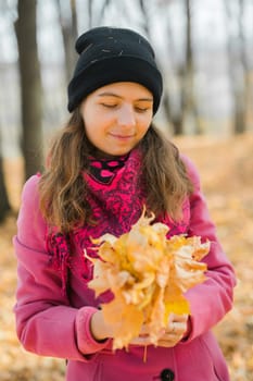 Portrait of beautiful happy young woman in pink jacket holding bouquet of yellow leaves at autumn park. Pretty Caucasian lady smiling and looking at camera during her walk outdoors. Generation Z and gen z youth.