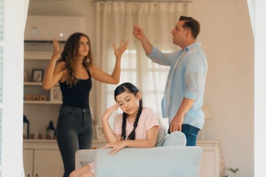 Annoyed and unhappy young girl sitting on sofa trapped in middle of tension by her parent argument in living room. Unhealthy domestic lifestyle and traumatic childhood develop to depression.Synchronos