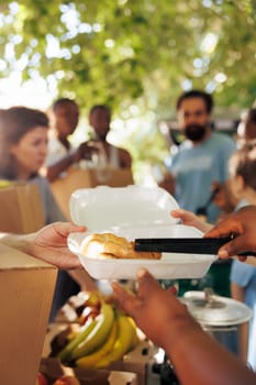 Photo focus on african american volunteer serving warm meal to needy hungry caucasian individual at a food drive. Detailed shot of poor less privileged person receiving free food from charity worker.