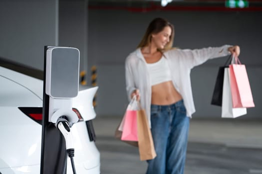 Young woman travel with EV electric car to shopping center parking lot charging in downtown city showing urban sustainability lifestyle by green clean rechargeable energy of electric vehicle innards