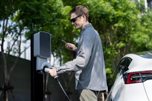 Young man travel with EV electric car charging in green sustainable city outdoor garden in summer shows urban sustainability lifestyle by green clean rechargeable energy of electric vehicle innards