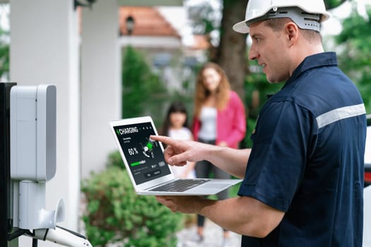 Qualified technician working on home EV charging station installation, making troubleshooting and configuration setup on charging system with laptop for EV at home with the family present. Synchronos