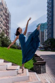 Beautiful Asian ballerina in a blue dress stands on the stairs in the splits outdoors. Urban landscape. Vertical photo