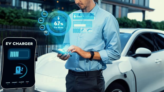 Businessman checking battery status hologram from EV smartphone application while his EV car recharging from charging station in residential area. Futuristic clean sustainable EV tech. Peruse