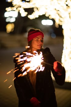 Woman holding sparkler night while celebrating Christmas outside. Dressed in a fur coat and a red headband. Blurred christmas decorations in the background. Selective focus.