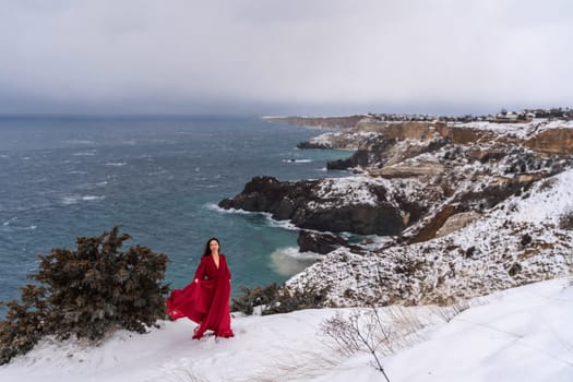 Woman red dress snow sea. Happy woman in a red dress in the snowy mountains by the emerald sea. The wind blows her clothes, posing against sea and snow background