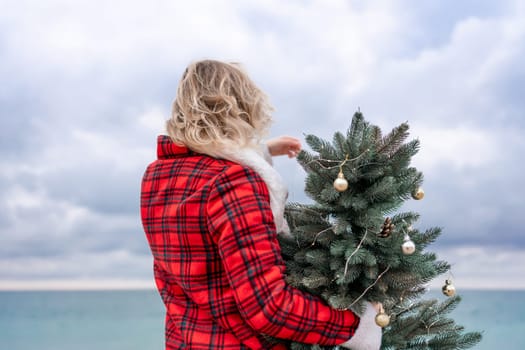 Blond woman holding Christmas tree by the sea. Christmas portrait of a happy woman walking along the beach and holding a Christmas tree in her hands. Dressed in a red coat, white suit