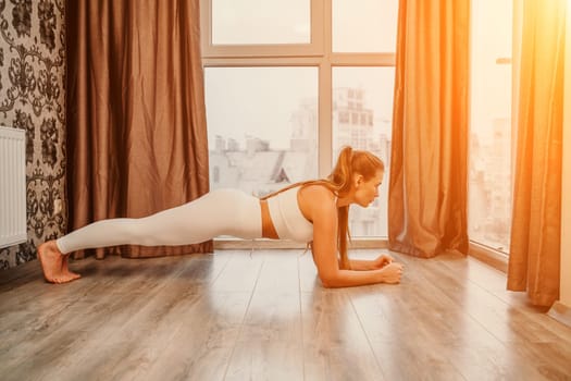 Young attractive athletic woman in white sportswear doing push ups or pushups on gym mat at home, practicing yoga plank pose, exercising indoors, healthy active lifestyle concept.