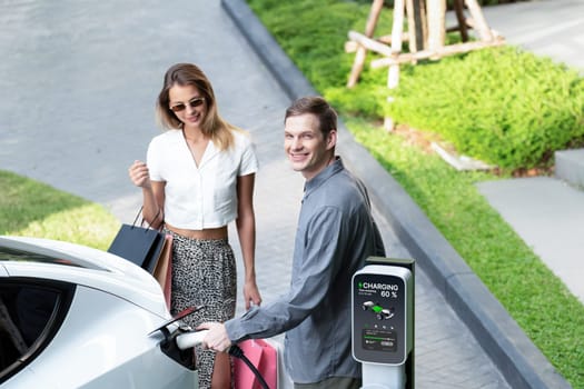 Young couple travel with EV electric car charging in green sustainable city outdoor garden in summer shows urban sustainability lifestyle by green clean rechargeable energy of electric vehicle innards