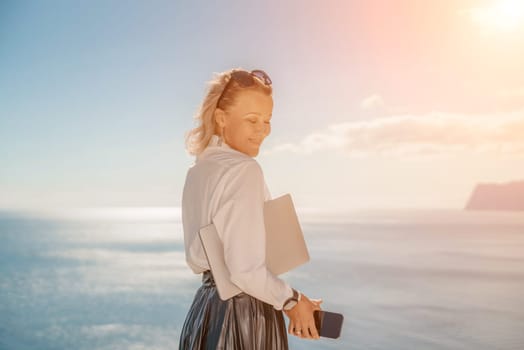 Business woman on nature in white shirt and black skirt. She works with an iPad in the open air with a beautiful view of the sea. The concept of remote work