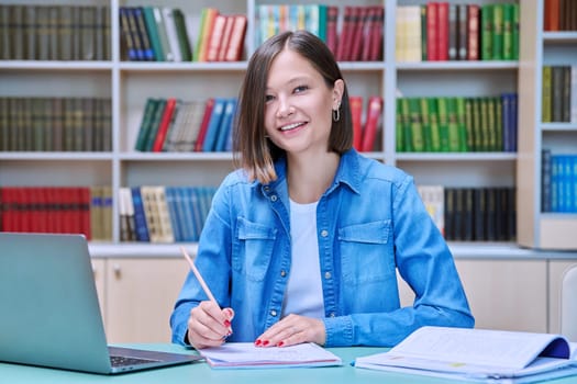 Portrait of smiling young female university student looking at camera sitting at desk with laptop in library classroom of educational building. Knowledge, higher education, youth concept