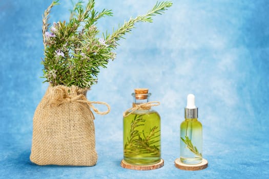 glass bottle and dropper with rosemary essential oil with fresh branches inside next to a raffia bag with freshly cut flowering rosemary branches on a blue background