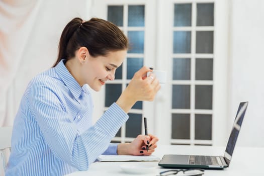 woman at a computer with a mug writes in a notebook in the office at a remote work Internet communication IT work