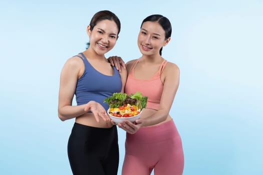 Two young sporty Asian women in sportswear holding salad bowl fill with fruit and vegetable. Natural youthful and fit body lifestyle people with balance nutrition on isolated background. Vigorous