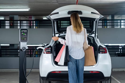 Young woman travel with EV electric car to shopping center parking lot charging in downtown city showing urban sustainability lifestyle by green clean rechargeable energy of electric vehicle innards