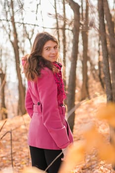 Cheerful young woman with pink scarf relaxing at park with yellow trees in background. Smiling beautiful girl enjoying warm sunny weather in autumn season. Happy pretty natural woman laughing in fall season