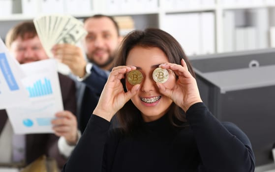 Young happy smiling chinese woman holding two coins of bitcoins in hand instead of glasses rejoices that she had time to buy to produce the moment deshego before the rise in price and growth.