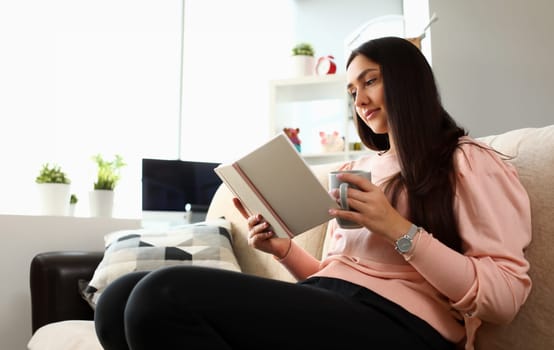 Young attractive indian teenager girl look into open book drink tea lying at couch portrait. Beautiful woman on sofa use datebook synopsis exam preparation delivery concept