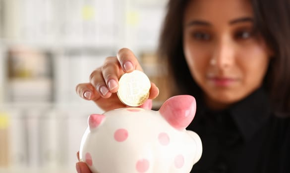 A young smiling chinese woman holding bitcoin coin in his hand thrusts it into piggy bank form of pink piggy saving storage theme in crypto currency.