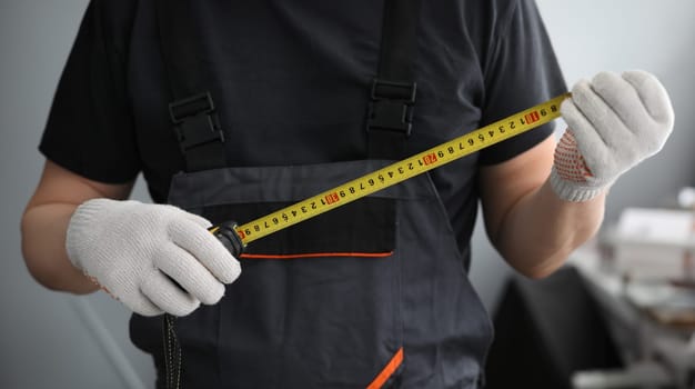 Focus on hands of handyman with roulette in hands. Tape for getting precise sizes. Person wearing special uniform and protective gloves at job. Toolkit of foreman