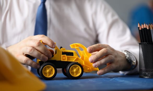 Close-up of businessman hands driving toy yellow excavator. Builder holding backhoe loader and showing future restructuring of big city. Business and construction concept