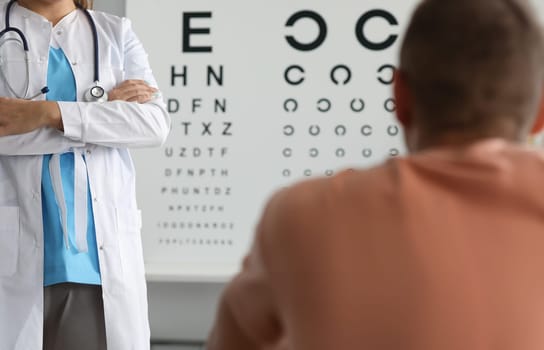 Focus on female ophthalmologist standing in clinic office and performing health examination to male patient. Oculist crossed hands on chest and waiting for result. Healthcare and medicine concept