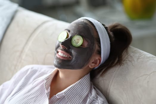 Woman makes gray clay facial mask at home. Put cut cucumber on her eyes like glasses. Spa home beaity skin mask make concept