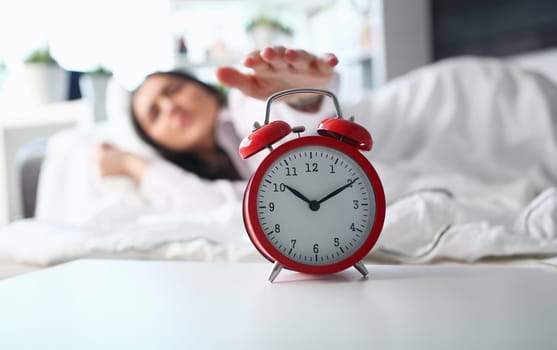 Sleepy young woman portrait with one opened eye trying kill alarm clock. Early wake up not getting enough sleep going work concept. Female stretching hand to ringing alarm willing turn it off