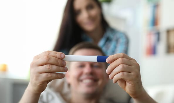 Focus on husband with ring on finger holding hcg checker and smiling. Happy wife on background. People ready to become parents. Pregnant woman waiting baby