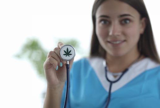 Focus on female hand holding stethoscope with cannabis. Smiling doctor wearing white gown and showing medical instrument with marijuana sign on it. Medicine and healthcare concept