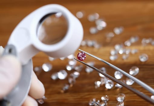 Goldsith in white gloves hold tweezers with pink gemstone and magnifier lens closeup background. Assessment of quality diamonds concept