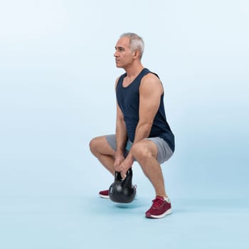 Full body length shot athletic and sporty senior man doing squat with kettlebell for body workout on isolated background. Healthy active physique and body care lifestyle after retirement. Clout