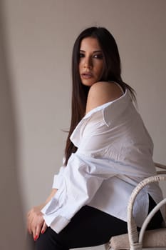 fashionable woman in a white shirt sits in a chair on a light background in the room