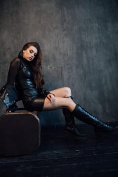 fashionable brunette in black leather clothes on a chair on gray background in the room