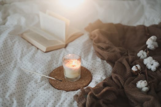 Cozy still life interior details with a book, candle and a cotton flower in warm soft bed. Sweet home