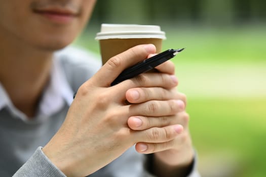 Closeup shot of man hand holding paper cup of coffee, sitting in the public park.
