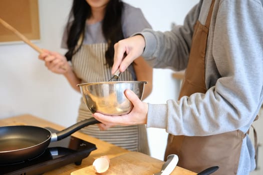 Young man wearing apron mixing ingredients in bowl, preparing breakfast with girlfriend in kitchen.