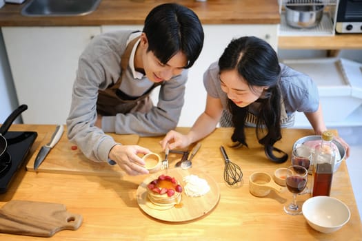 Joyful young couple putting honey on delicious pancakes with strawberries, preparing breakfast in cozy kitchen.