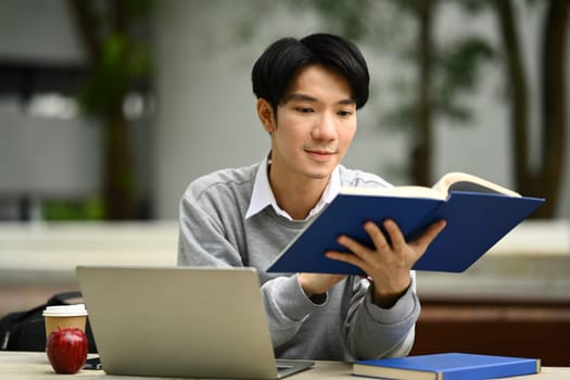 Peaceful young asian man student reading interesting book while sitting bench in sunny summer park.