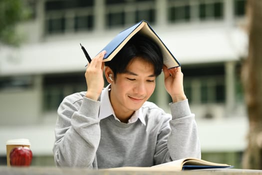 Happy asian student man putting book on his head, preparing for exam in campus. Education, Learning and lifestyle.