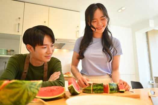 Pretty young woman and boyfriend cutting watermelon on wooden kitchen tabletop.