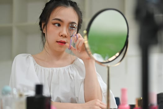 Beautiful young woman using eyelash curler curling eyelashes before mascara in front of mirror.