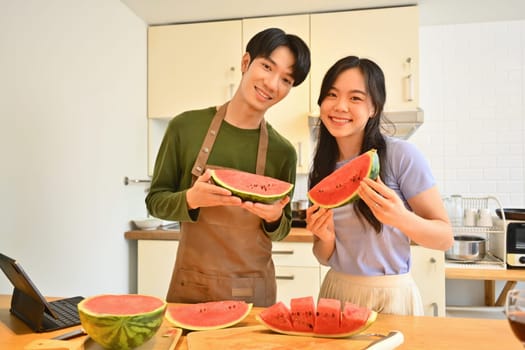 Happy young couple holding and eating slices of watermelon in home kitchen. Family love and casual lifestyle.