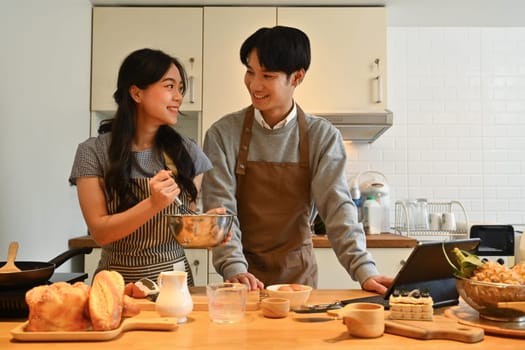 Cheerful young couple preparing breakfast in the kitchen at home and reading recipes on digital tablet.